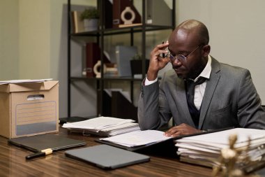 Gözlüklü siyah bir adam masasında oturmuş evrakları inceliyor ve not alıyor. Etrafı evrak ve ofis malzemeleriyle çevrili.