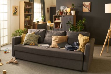 Dağınık oyuncaklarla dolu gri kanepe, dizüstü bilgisayar, sırt çantası ve kitapların yer aldığı modern oturma odası, arka planda çocuk veya gencin, bitki ve rafların olduğunu gösteriyor.