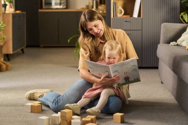 Kafkasyalı genç bir kadın, ahşap bloklarla çevrili, hikayeye odaklanmış, rahat ev ortamında, beyaz bir kıza resim kitabı okuyor.