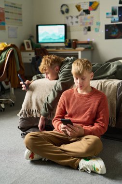 İki Kafkas genci yatak odasında akıllı telefon kullanıyor, biri yere oturmuş cihaza odaklanmış, diğeri ekrana bakıyor, arka planda görünen modern iç mekan.