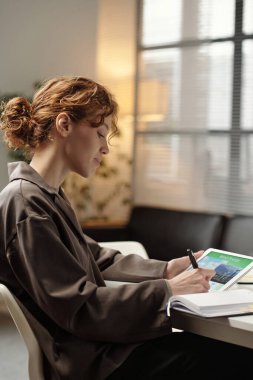 Z Geni beyaz kadın ofis masasında oturuyor, dijital tablet ekranına bakıyor ve not defterine yazıyor, dikey yan görünüm