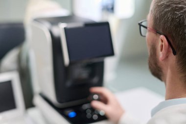 Klinik ortamda göz muayenesi sırasında tıbbi ekipmanların önünde otururken kontrol düğmesi kullanan beyaz, orta yaşlı bir adam oftalmik tanı cihazı kullanıyor.