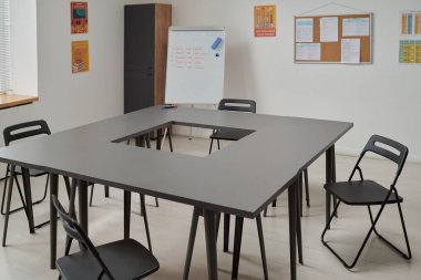 Boş sınıflar, masaların kare düzeniyle ve el yazısıyla yazılmış notları gösteren siyah sandalyelerle duvarlarda asılı eğitim posterleri ve çizelgeleri, pencereye giren güneş ışığıyla dolu.