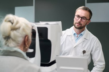 Kafkas kıdemli kadın göz muayenesinden geçerken modern tıp kliniğinde beyaz yetişkin erkek optometrist görme testi yapıyor.