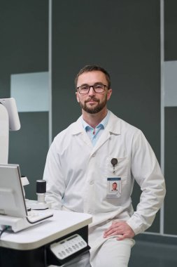 Klinik ortamda laboratuvar önlüğü giyen beyaz bir adamın portresi. Kucağında el ele tutuşan tarafsız bir ifadeyle kameraya bakıyor.