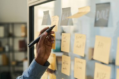 Cam duvara yapıştırılmış yapışkan notlar yazan siyahi adam, görevleri düzenleyen ve proje akışını planlayan, el ele tutuşan, işle ilgili çeşitli hatırlatmalar ve hedefleri olan görünür notlar.