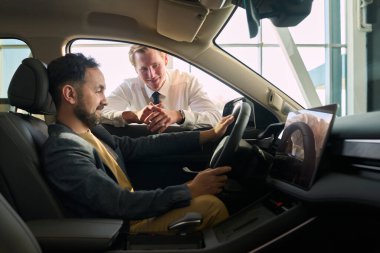 Beyaz, yetişkin bir adam şoför koltuğunda otururken beyaz, orta yaşlı bir adam arabanın penceresinde dikiliyor.