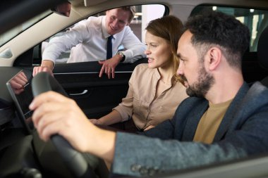 Beyaz, yetişkin bir adam sürücü koltuğunda elinde direksiyon tutarken beyaz, genç bir kadın gösterge panelini inceliyor.