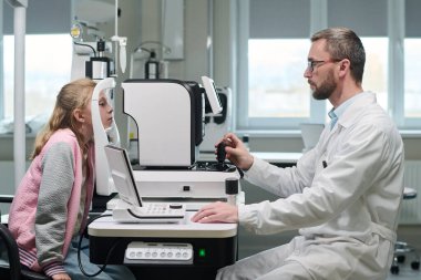 Klinikte tıbbi malzemeler kullanan göz doktoruyla göz muayenesine giren beyaz bir kız çocuğu, görme testi sırasında beyaz bir erkek doktor teşhis cihazı kullanıyor.
