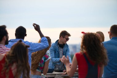 Çeşitli genç yetişkinler dans ederken Kafkasyalı genç adam çatıda DJ 'lik yapıyor, insanlar enerjik jestlerle DJ' e bakıyor, şehir manzarası arka planda görünüyor.