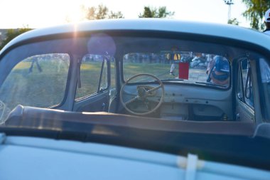 Arabanın içi boş ön koltukları ve arka pencereden içeri güneş ışığı süzülen klasik direksiyonu arka planda insanlar ve ağaçlarla açık hava ayarlarını gösteriyor.
