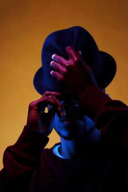 Fötr şapka takan genç siyahi bir erkeğin portresi. Stüdyoda soluk bir ışıkla poz verirken, gözleri görünmeden.