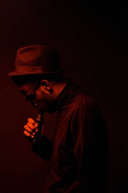 Gözlüklü ve fötr şapkalı şık Afro-Amerikan bir adamın portresi. Karanlık odada kırmızı ışıklı elektronik sigara buharlaştırıyor.