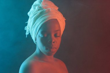 Geleneksel baş örtüsüyle çıplak omuzlarıyla ve koyu arkaplanda kapalı gözlerle poz veren güzel bir Afrikalı kadının belden yukarı portresi.