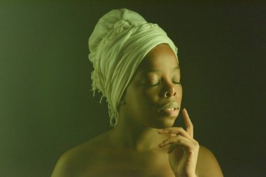 Çekici, çıplak omuzlu, beyaz sarıklı, gözleri koyu arkaplanda poz veren siyah bir kadının yakın plan portresi.