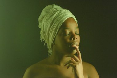 Belden yukarısında, omuzları açık, beyaz başlıklı Afro-Amerikan bir kadının koyu arkaplanda gözleri ve dudakları kapalı poz verişi.