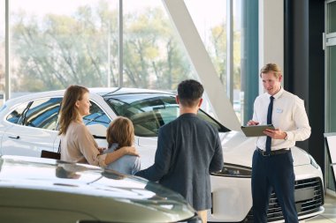 Beyaz erkek araba satıcısı elinde dosyayla beyaz bir adama, beyaz bir kadına ve yeni araçların yanındaki galeride beyaz bir çocuğa yardım ediyor.