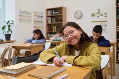 Asyalı genç kızın portresi, arka planda okuyan diğer gençlerle birlikte sınıfta otururken, sıralarda kitap ve okul gereçleri okurken ve deftere gülümseyip yazarken.