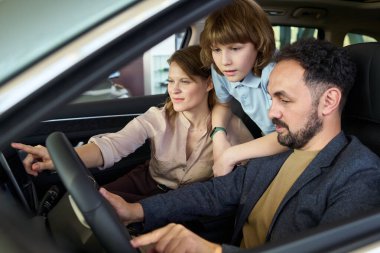 Beyaz, orta yaşlı bir adam sürücü koltuğunda oturuyor ve modern araba galerisindeki kadın ve çocuğa araba kontrolü yapıyor.