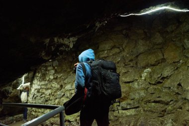 Sırt çantalı genç adam yeraltı mağarasını keşfediyor, metal parmaklıklarla taş duvar boyunca yürüyor, başka bir orta yaşlı beyaz adam arka planda dikilip çevreyi gözlüyor.