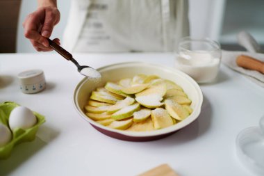 Bilinmeyen bir kadın fırın kabına elma serpiyor. Mutfakta tatlı hazırlıyor. Tezgahta şekerli yumurta ve cam kâse var.