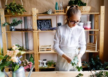 Çiçekçi dükkanında yeşil yaprakları düzenleyen, gözlük ve önlük takan, çiçek malzemeleri ve süslemelerle ahşap rafların önünde duran beyaz bir kadın.