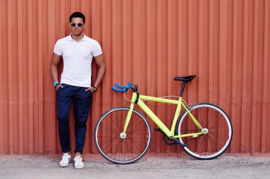 Sarı yol bisikletinin yanında kolları çapraz duran genç Latin adam turuncu bir duvara yaslanmış, güneş gözlüğü takıyor ve kameraya güvenle bakıyor.