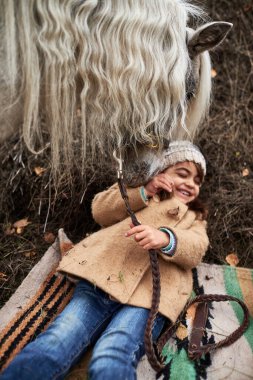 Beyaz bir kız çocuğu yerde yatarken gülümsüyor at dizginlerini tutarken, at burnunu sokuyor, açık havada sonbahar ayini düzenliyor, genç kız ve hayvan arasında neşeli bir etkileşim var.
