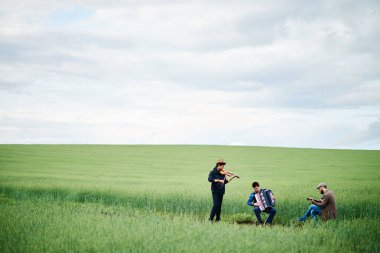 Üç yetişkin adam yeşil alanda müzik enstrümanları çalıyor, biri keman çalıyor, biri akordeon çalıyor, biri gitar çalıyor, ayakta duruyor ve açık kırsalda oturuyor.