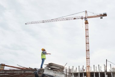 Kafkas kasklı orta yaşlı bir adam inşaat alanında dikiliyor ve inşaat malzemeleri ve tamamlanmamış yapılarla çevrili vinç kulesine bakıyor.