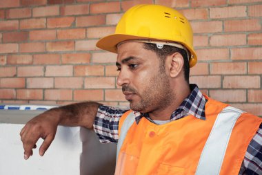 Güney Asyalı adam sıkı şapka ve güvenlik yeleği giyiyor inşaat malzemelerine yaslanıyor, ciddi bir ifadeyle kafasını çeviriyor, tuğla duvarın önünde duruyor.