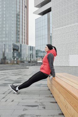 Soğuk bir sonbahar sabahı, iş bölgesinde ahşap bankta fiziksel egzersiz yapan genç atletik beyaz kadının yan görüntüsü.