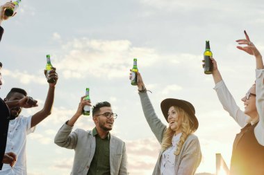 Erkek ve kız öğrencilerden oluşan çok ırklı bir grup çatıdaki terasta takılırken bira ve dans şişelerini kaldırıyorlar.