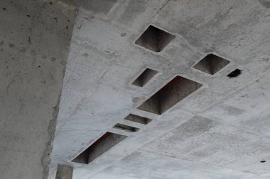 Beton tavan birden fazla dikdörtgen kesikleri ve tamamlanmamış yüzeyi gösteriyor, açıkta kalan yapısal elementler ve hammaddedeki geometrik açıklıkları içeren inşaat alanı
