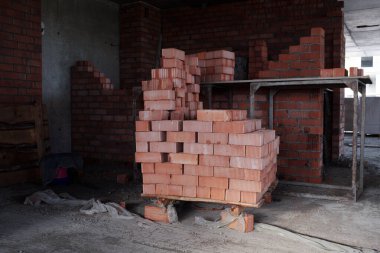 Ahşap paletin üzerine yerleştirilmiş kırmızı tuğla yığını inşaatı tamamlanmamış binanın içinde duruyor, tuğla duvarlar ve arka planda beton zemin görünüyor, kimse yok.