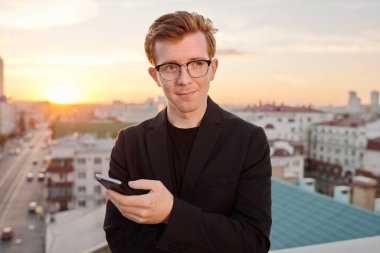 Genç, çekici, kızıl saçlı, beyaz bir adamın belden yukarı portresi. Çatıda durmuş, akıllı telefonuyla dikkatle başka tarafa bakıyor.