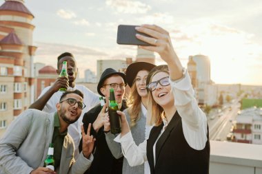 Young pretty woman taking selfie on smartphone with her stylish cheerful friends hanging out on rooftop terrace with beer at sunset