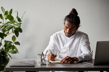 Ofis odasında oturan şık beyaz gömlek ve şık aksesuarlar giyen Hintli genç bir adamın yatay portresi not alıyor.