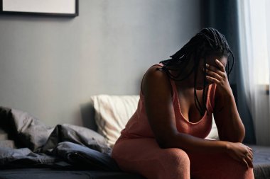 Genç siyahi bir kadının depresyonda olduğunu gösteren yatay bir fotoğraf. Yatak odasında oturup, gözlerini kapayıp, sorunları düşünürken.