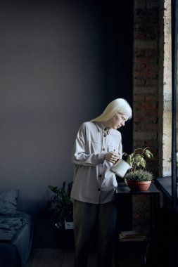 Modern albinizmli genç bir kadının dikey resmi. Gri bir kıyafet giyiyor. Çatı katında çiçekleri suluyor.