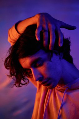 Dikey yakın plan portre çekimi. Yakışıklı genç adam şık giyinmiş. Mavi ve turuncu neon ışıkta gözleri kapalı kameraya poz veriyor.