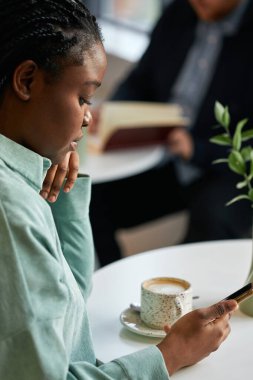Modern siyah bir kadının modern bir kafede kahve molası verip akıllı telefondan internette bir şeyler izlediği dikey bir fotoğraf.