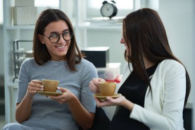 Kısa saçlı, gri elbiseli, yuvarlak gözlüklü genç ve şık bir kadın, kadın arkadaşıyla birlikte kanepede oturuyor. Küçük bej bardaklardan kahve içerken neşeyle konuşup gülümsüyorlar.