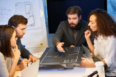 Deneyimli tasarımcılar ofiste masalarında oturmuş, ellerinde siyah dokuma kağıdından yapılmış yeni bir ev düzeni tutuyorlar. Sakallı şef taslağa işaret ediyor ve bazı düzeltmeler öneriyor.