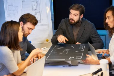 Genç erkek ve kadın uzmanlar ofis masasında oturmuş ortada cereyan yapıyorlar. Şık sakallı adam projeye işaret ediyor ve iş arkadaşlarıyla iç mimari fikirlerini paylaşıyor.