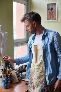 Sanat stüdyosunda çalışıyorum. Lekeli önlüklü dalgın erkek sanatçı şaheser yaratmak için mükemmel bir fırça seçiyor.