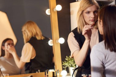 Profesyonel güzellik salonunda. Kadın modele makyaj yapan konsantre bir transseksüel makyöz büyük aynanın önünde sandalyede oturuyor.