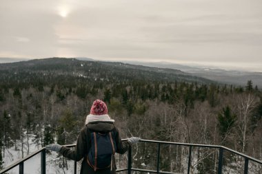 Üstü açık giyinen ve sırt çantalı bir kadın vadide bulutlu dağlara bakıyor.