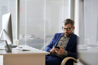 Kafkasyalı orta yaşlı sakallı yönetici modern ofiste dinleniyor, masada oturuyor ve akıllı telefondan mesaj atıyor.