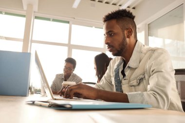 Modern ofiste dizüstü bilgisayarda çalışan genç siyahi bir adamın yan görüntüsü, arka planda konuşan bayan meslektaşları.